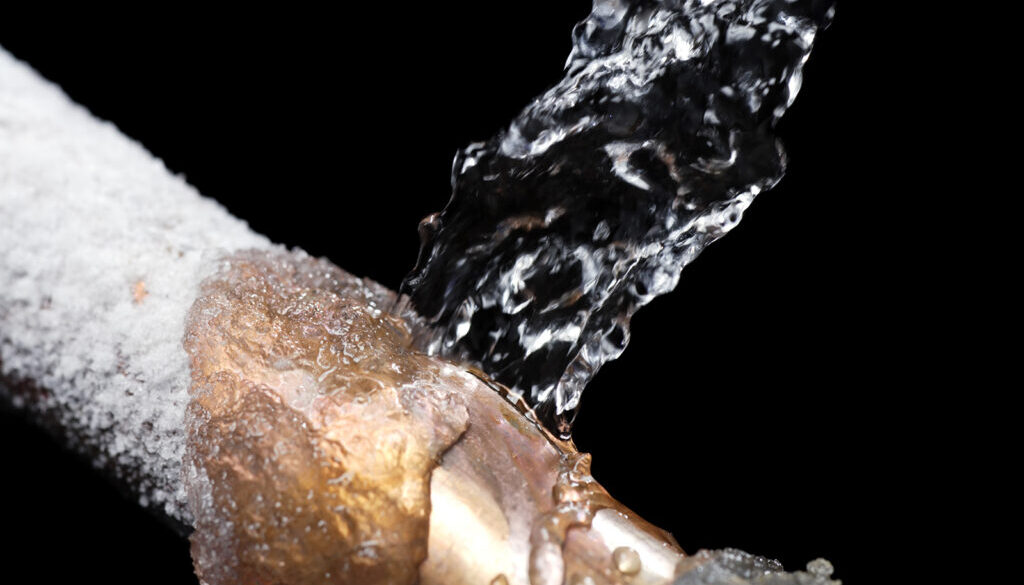 A copper water line set against a black backdrop. The copper line is seen close up with the top portion frozen and a small fractured crack where clear water is being expelled upward with force.